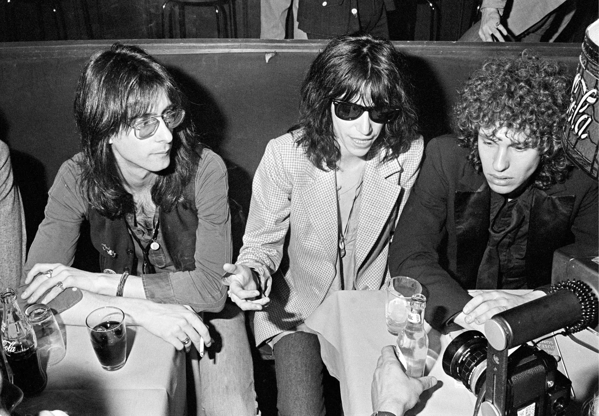 Three people with long hair sit at a table in a dimly lit bar or club, two wearing sunglasses. Drinks are on the table and a camera or recorder is visible in the foreground. The setting appears casual and candid.